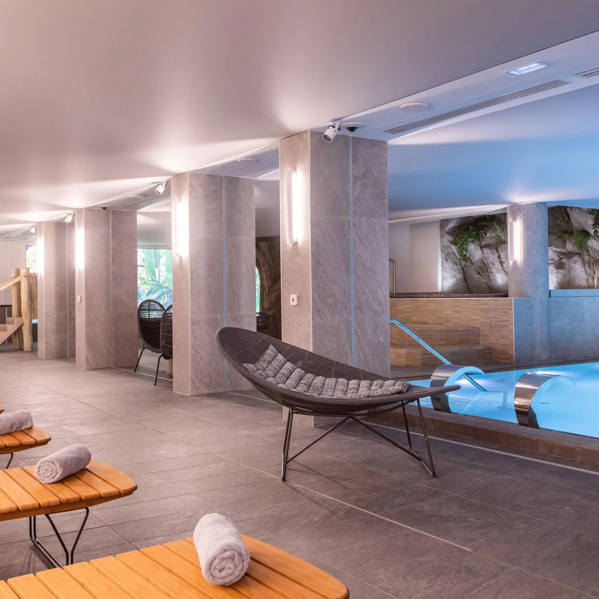 Salle de détente avec piscine d'eau bleue, chaises longues en bois et fauteuils, décorée de roches murales et de plantes