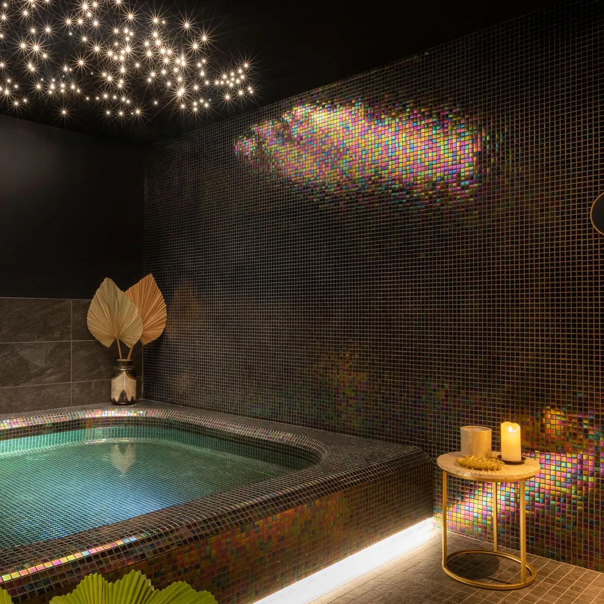 Baignoire encastrée dans un mur recouvert de mosaïque irisée, éclairée par des étoiles au plafond et une bougie sur une table basse