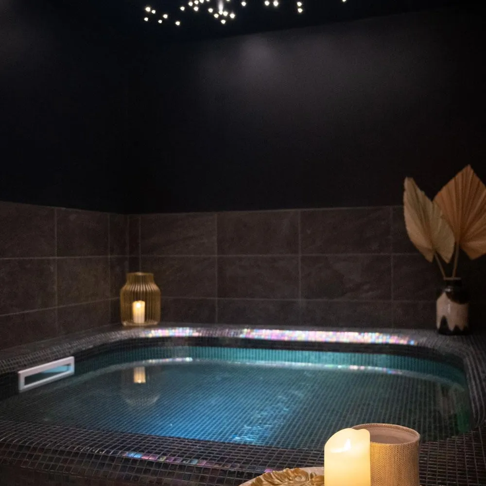 Piscine intérieure éclairée avec des lumières sous-marines, entourée de carreaux sombres, et une table avec une bougie allumée et un vase contenant une feuille décorative
