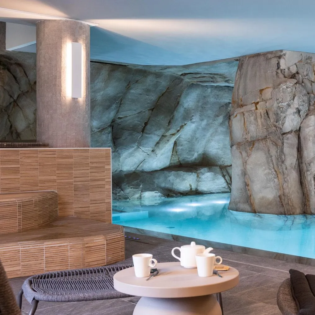 Un salon avec une table basse, des tasses et une théière, devant un mur décoré de motifs rocheux et une piscine éclairée