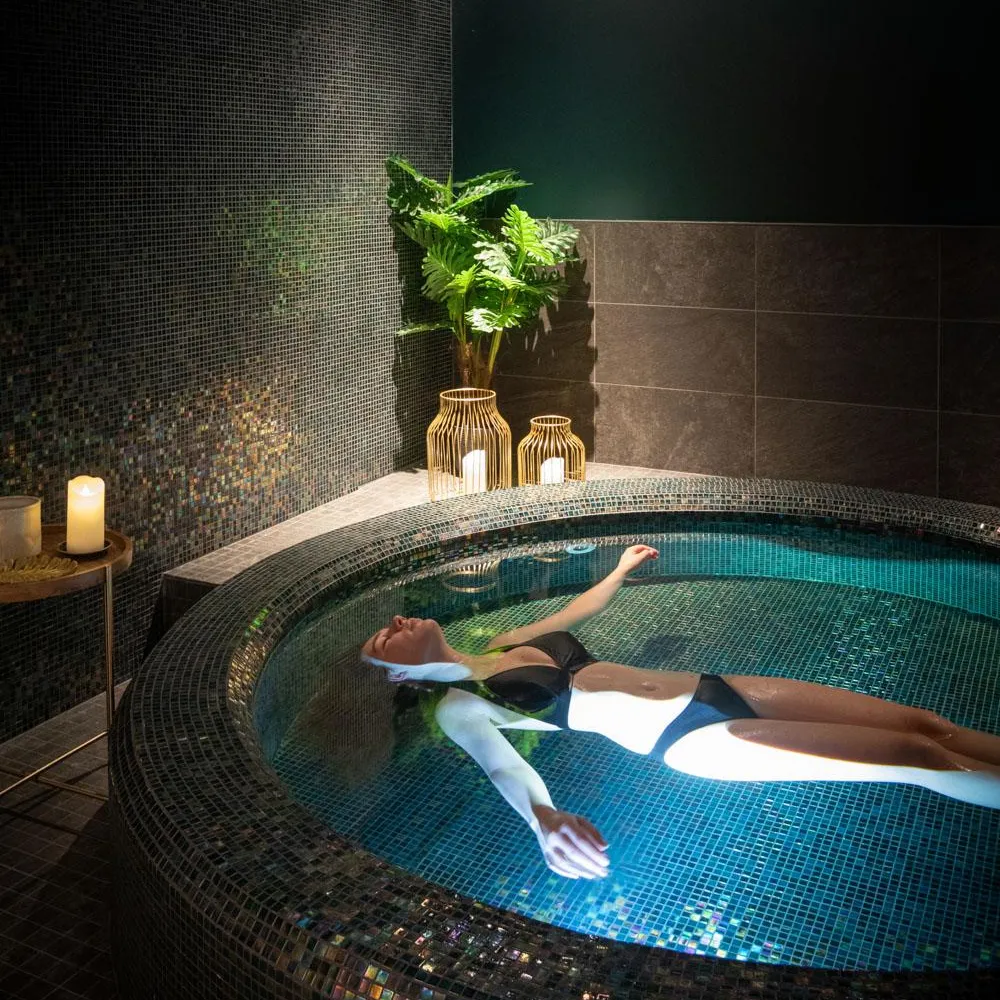 Une personne en maillot de bain flotte dans un bain à remous entouré de mosaïques, avec des bougies et des plantes en arrière-plan