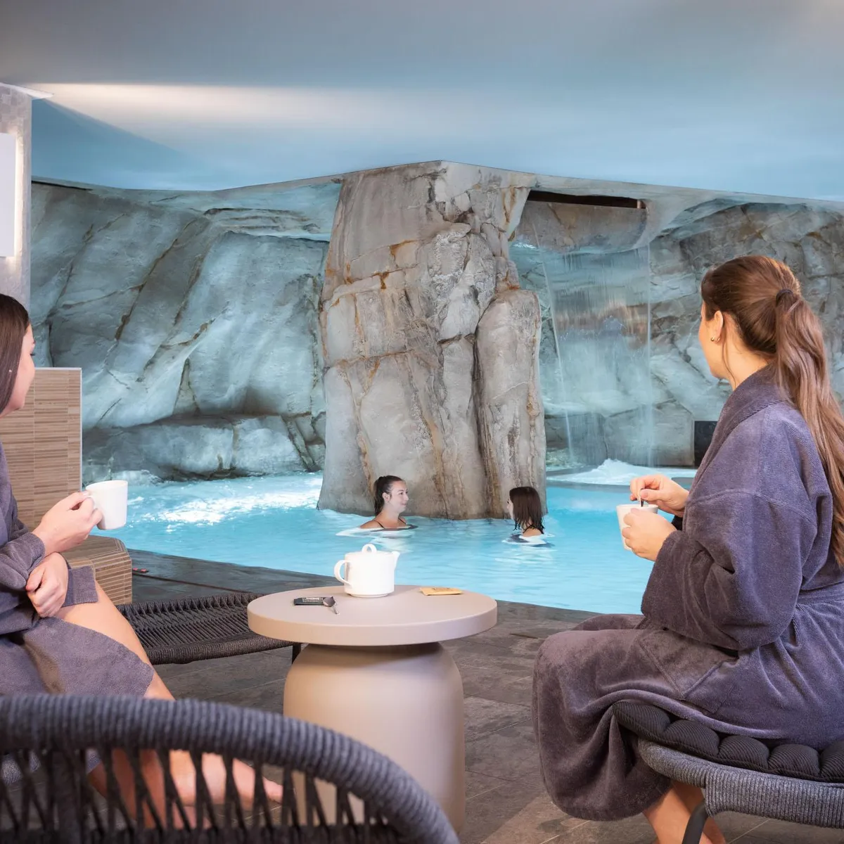 Deux personnes assises en robe de bain discutent près d'une piscine intérieure avec cascade et rochers artificiels, tenant des tasses, tandis que d'autres personnes nagent