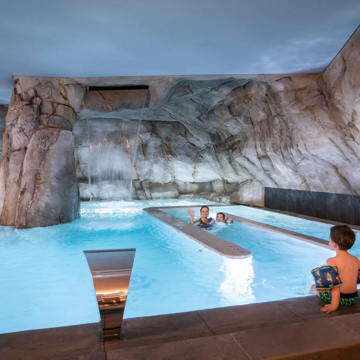 Une piscine intérieure avec cascade artificielle et plusieurs personnes nageant ou s'apprêtant à nager
