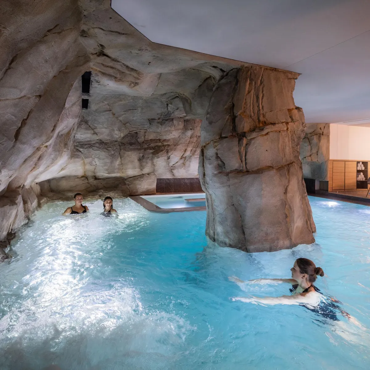 Trois personnes nagent dans une piscine intérieure aux parois rocheuses, avec un espace de détente visible en arrière-plan