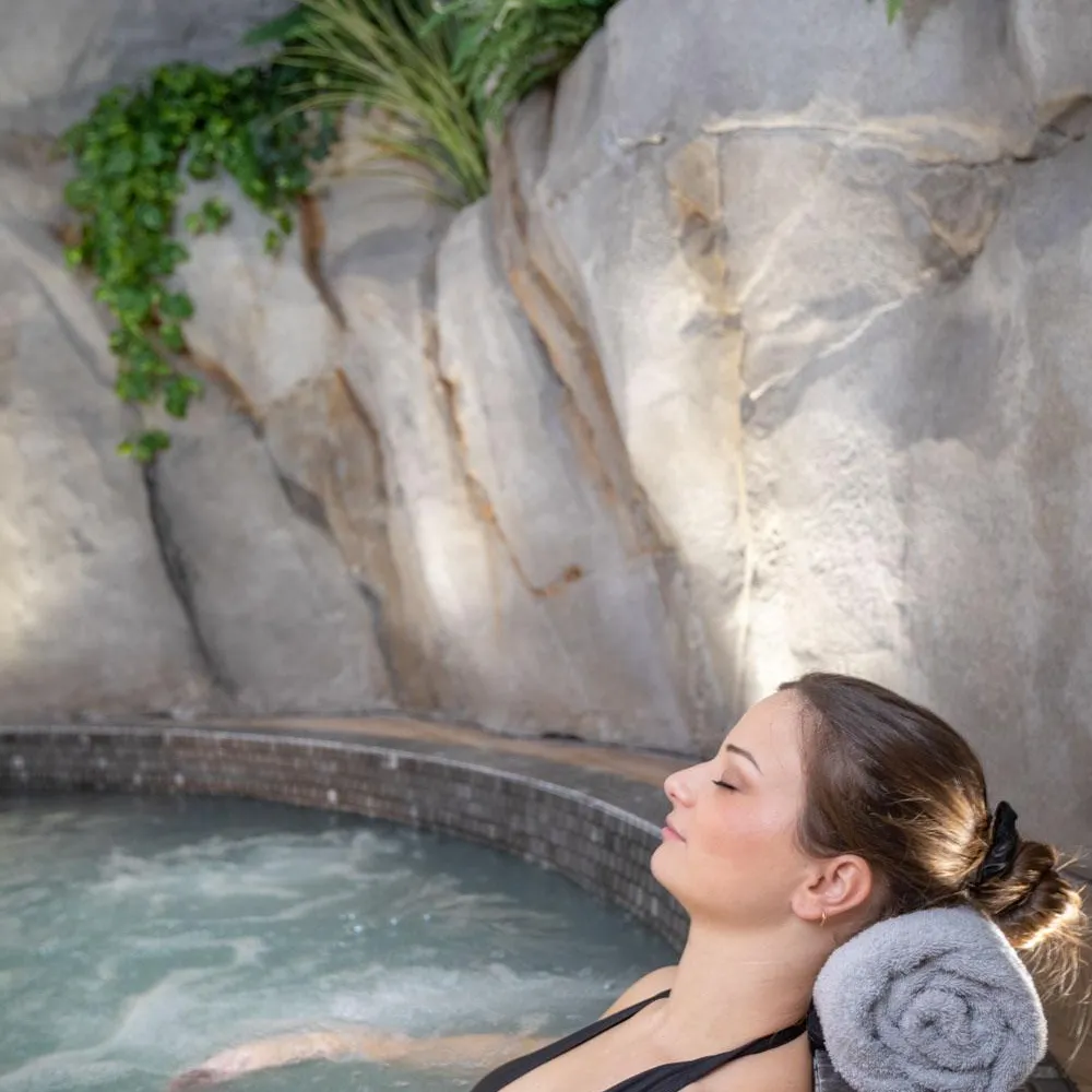 Femme en maillot noir allongée dans un bain à remous avec une serviette grise derrière la tête, entourée de rochers et de végétation