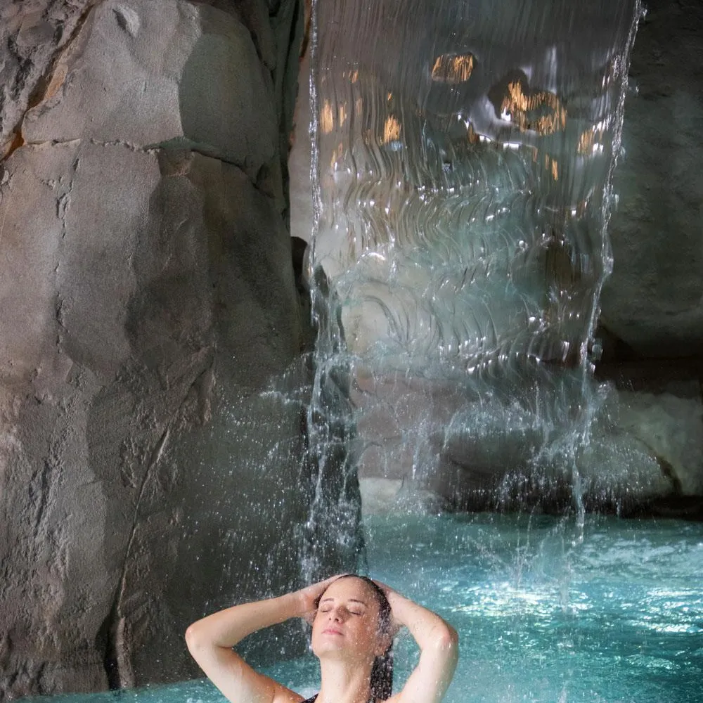 Une personne est immergée dans une piscine sous une cascade d'eau, les mains derrière la tête, dans un cadre rocheux