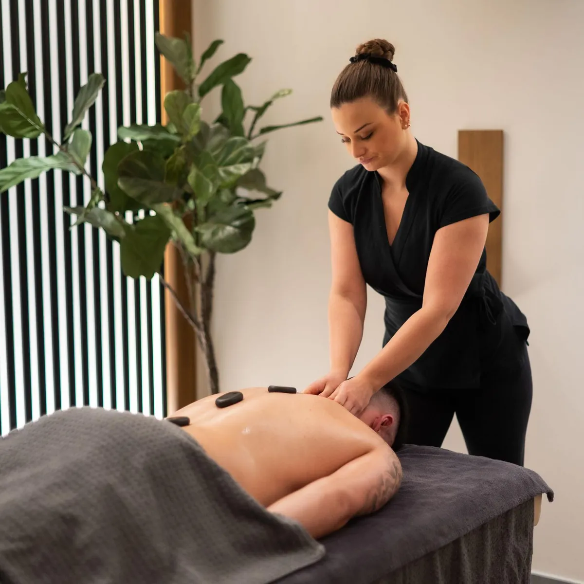 Une femme en tenue noire pratique un massage sur le dos d'une personne allongée sur une table recouverte d'une serviette, avec une plante verte à proximité