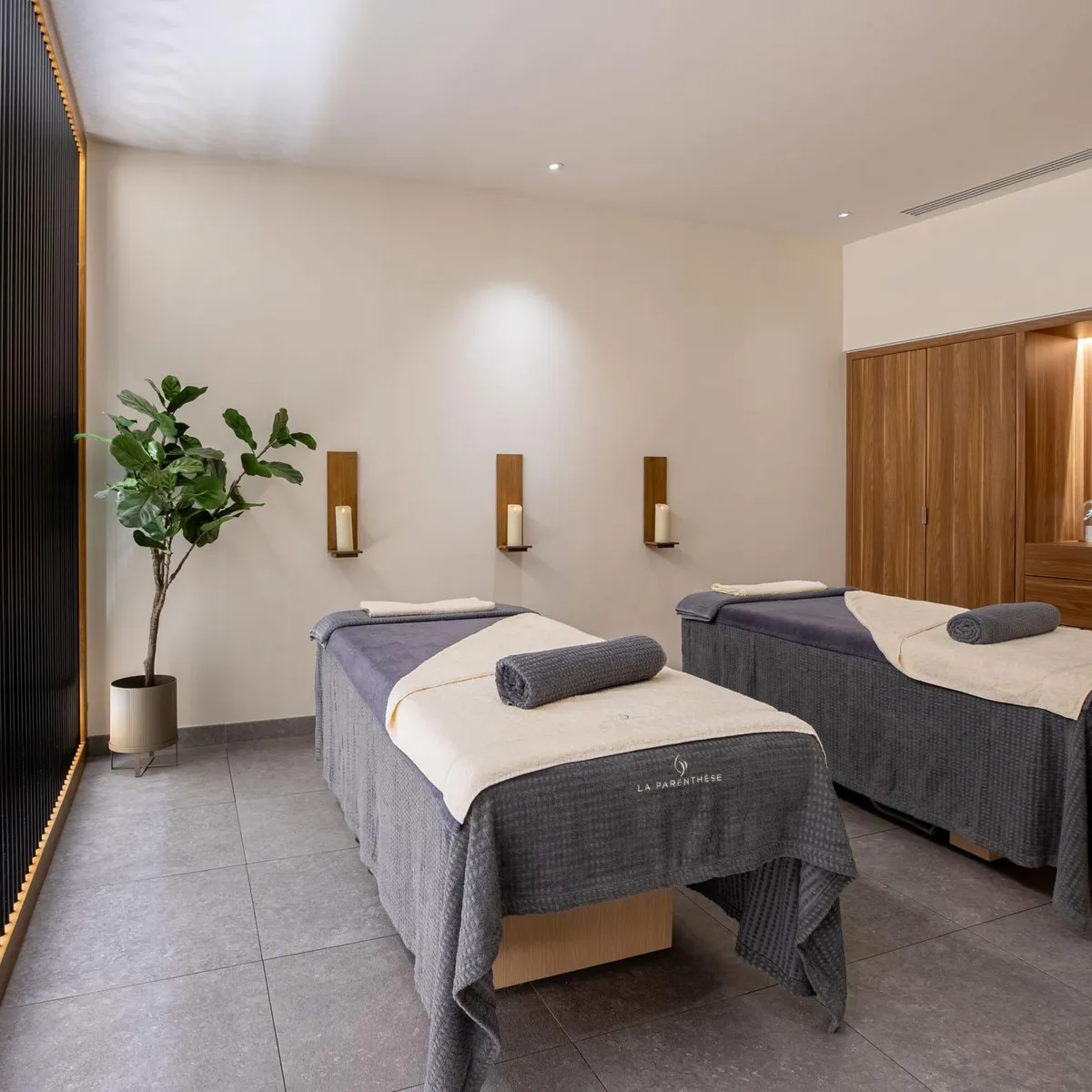 Deux tables de massage avec serviettes grises et blanches, une plante verte et des appliques murales dans une pièce aux murs beiges et au sol gris