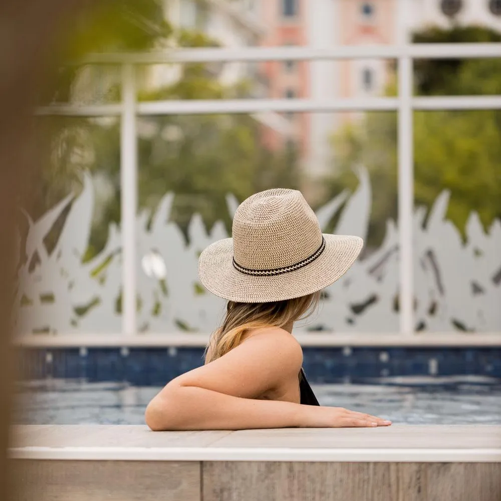 Une personne portant un chapeau de paille est assise au bord d'une piscine, le dos tourné vers le spectateur, avec un bâtiment en arrière-plan
