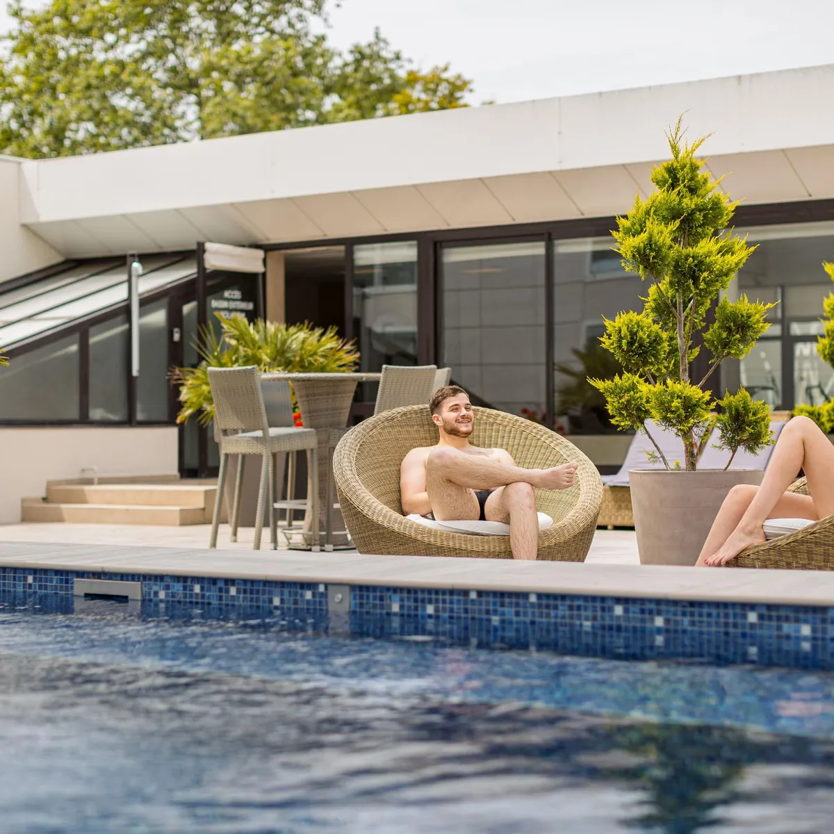 Deux personnes assises dans des fauteuils en osier au bord d'une piscine devant un bâtiment moderne avec des plantes en pot