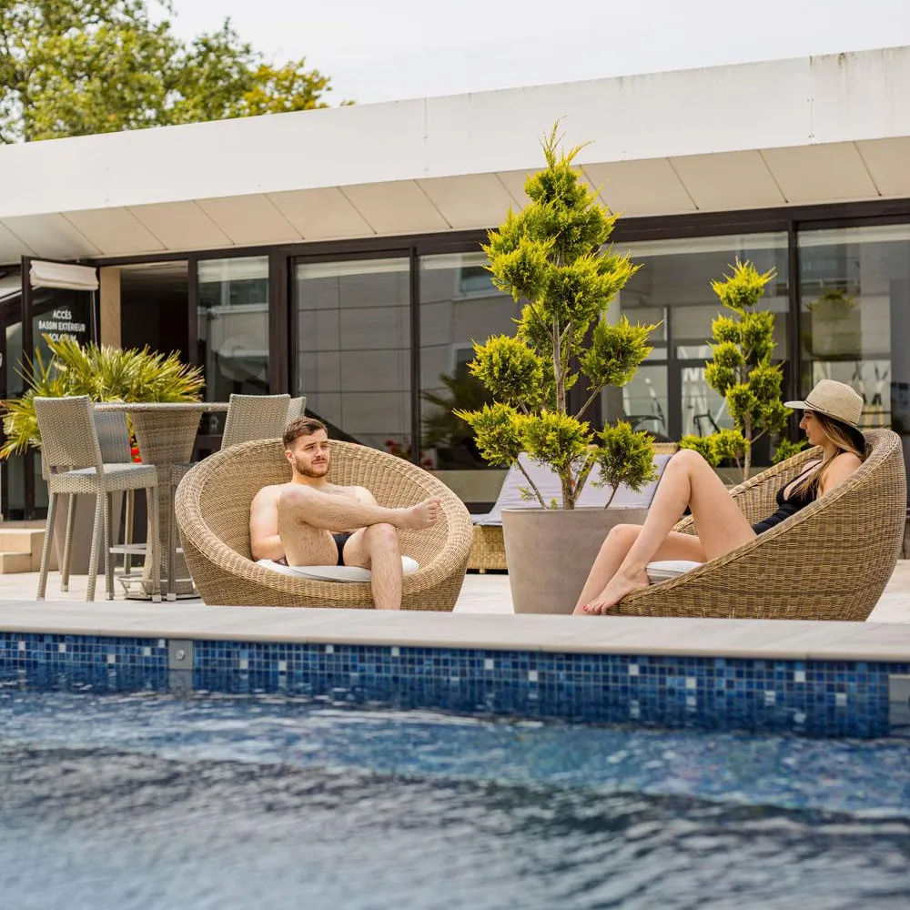 Deux personnes assises dans des fauteuils en osier au bord d'une piscine devant un bâtiment avec de grandes baies vitrées