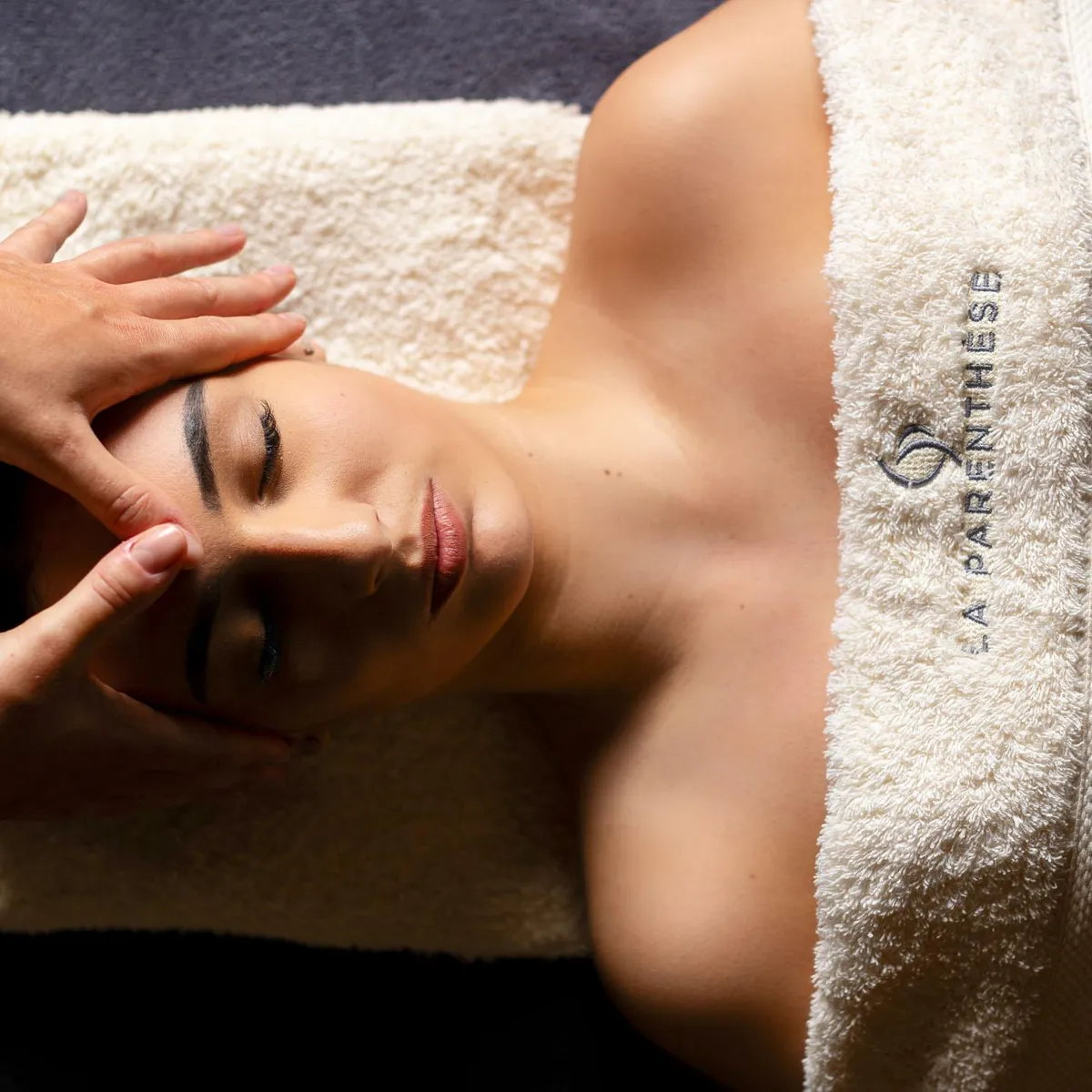 Une personne reçoit un massage facial, allongée sur le ventre, couverte d'une serviette avec une inscription