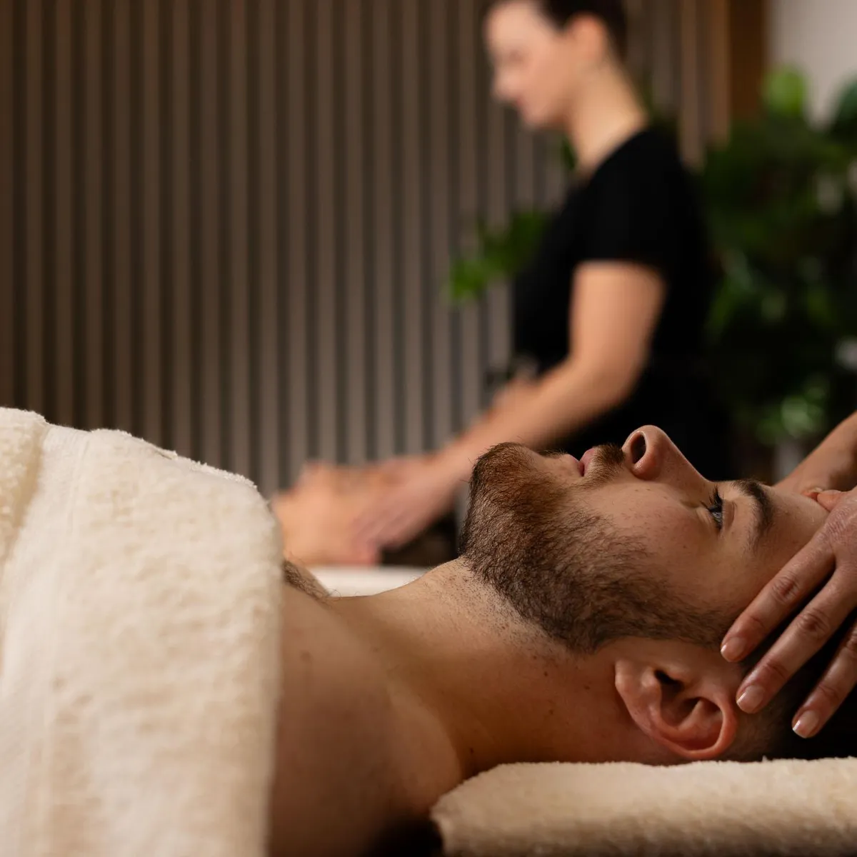 Un homme allongé sur le ventre reçoit un massage de la nuque par un professionnel, dans un cadre intérieur avec des plantes en arrière-plan