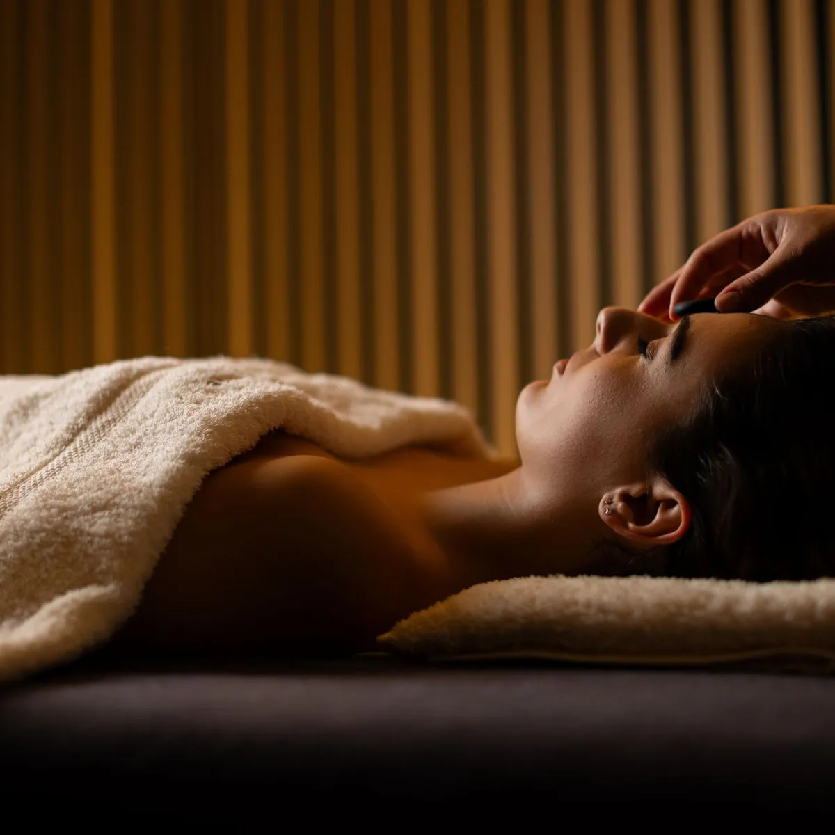 Une personne allongée sur une serviette reçoit un massage facial dans une pièce aux murs striés
