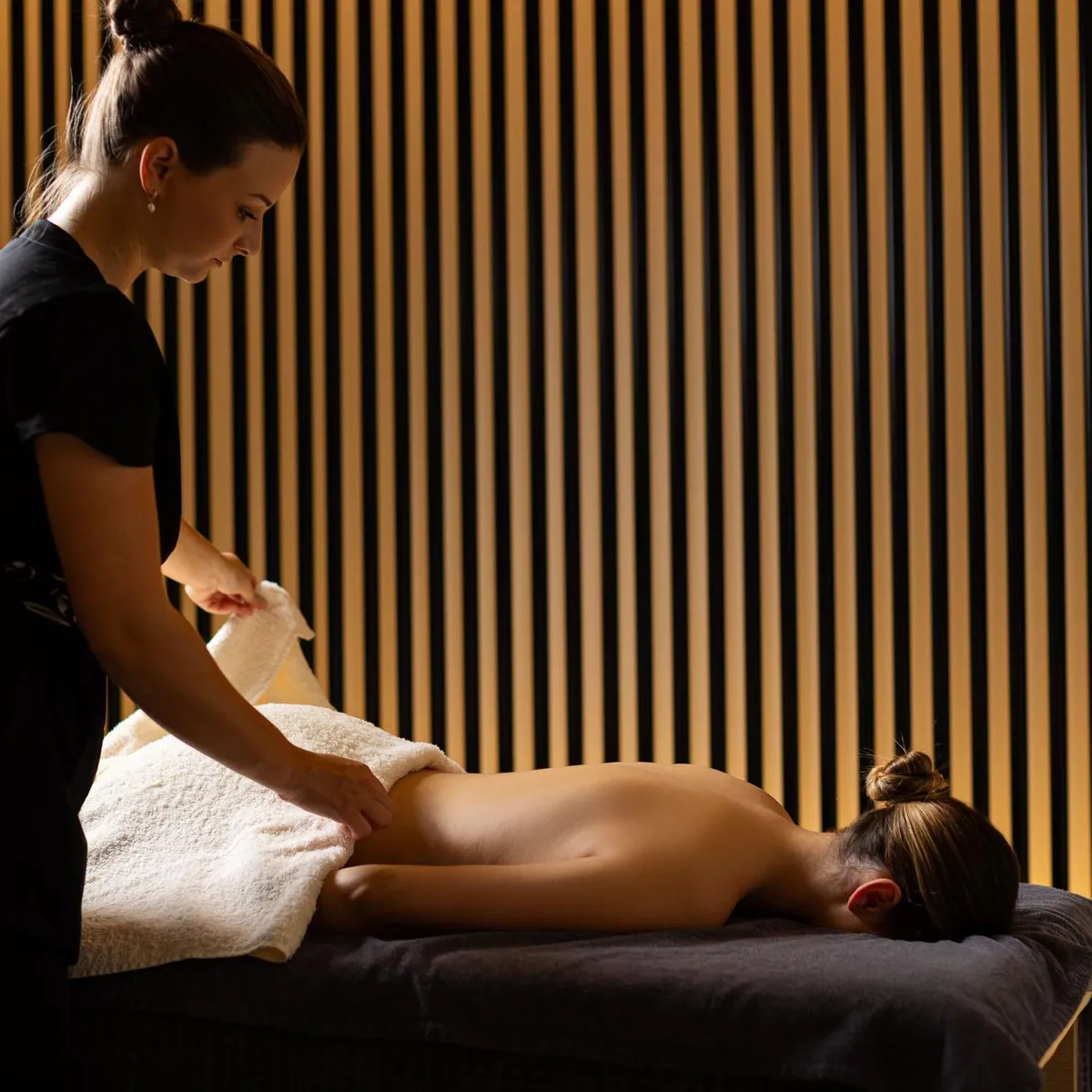 Une personne en tenue noire donne un massage à une autre personne allongée sur une table recouverte de serviettes