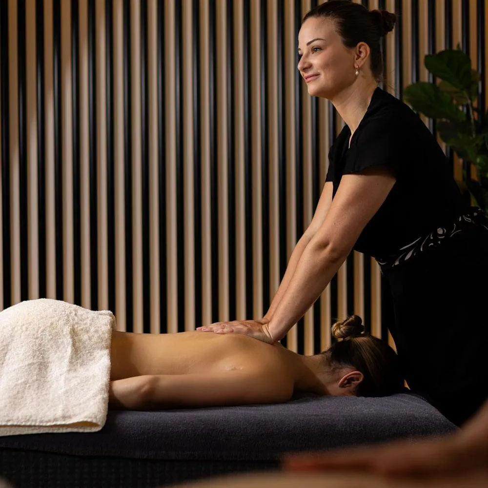 Une femme en tenue noire donne un massage à une personne allongée sur une table, dans une pièce aux murs à rayures verticales