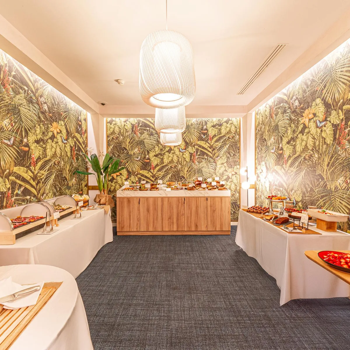 Buffet de restauration avec divers plats disposés sur des tables recouvertes de nappes blanches, dans une salle décorée de murs représentant une jungle tropicale