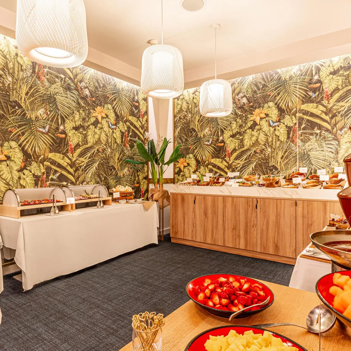 Buffet avec divers plats, dont une fontaine à chocolat, dans une salle décorée de motifs tropicaux et éclairée par des suspensions