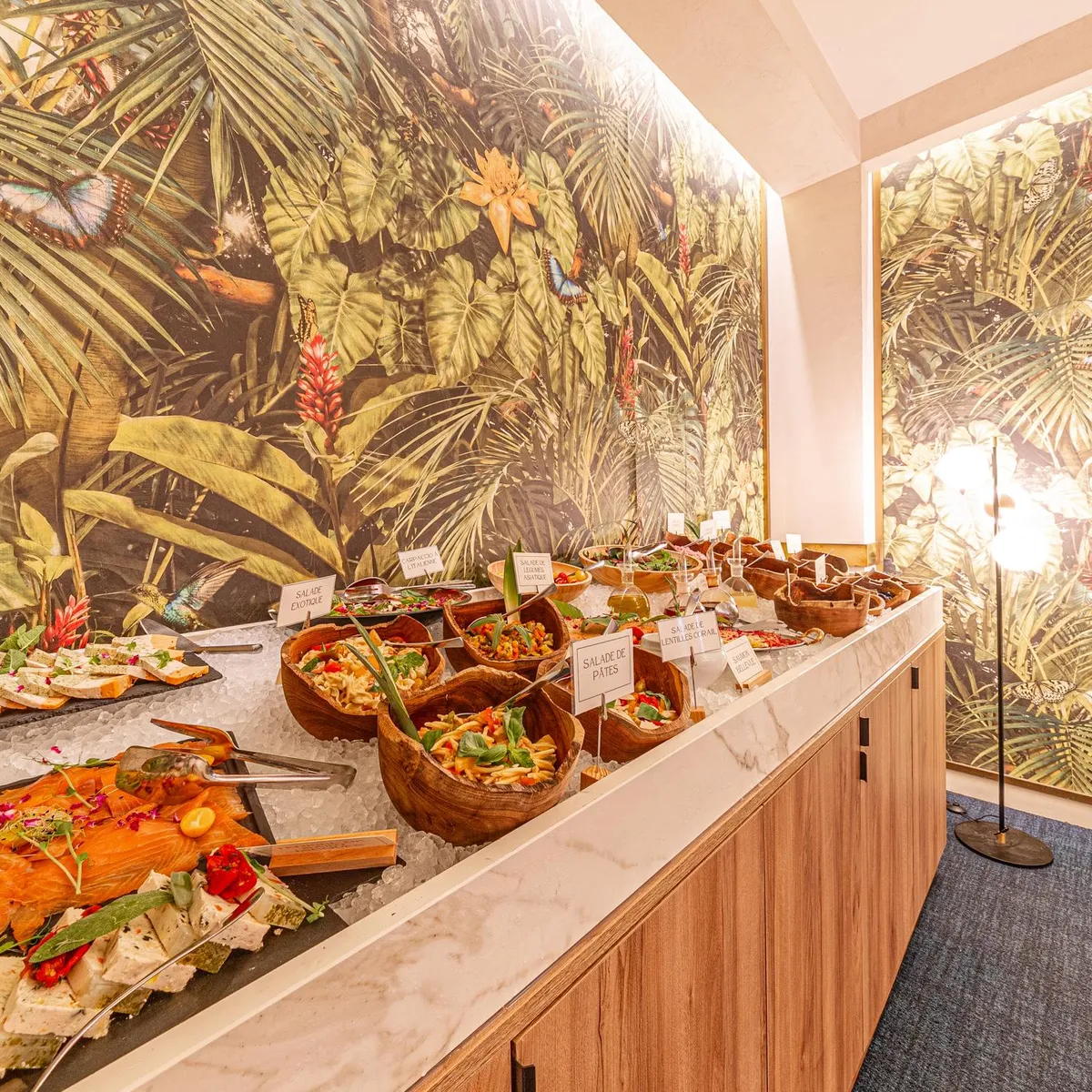 Un buffet de plats variés, dont du saumon fumé et des salades, est présenté sur une table recouverte de glace, avec des murs décorés d'un motif de feuilles tropicales