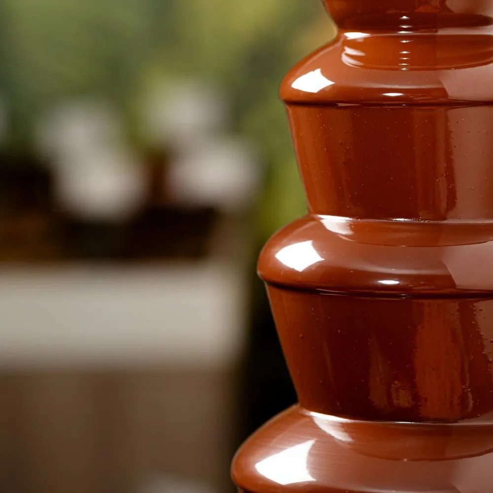 Un pot en céramique rouge à plusieurs niveaux est placé à l'extérieur devant un fond flou de verdure et de structures en pierre
