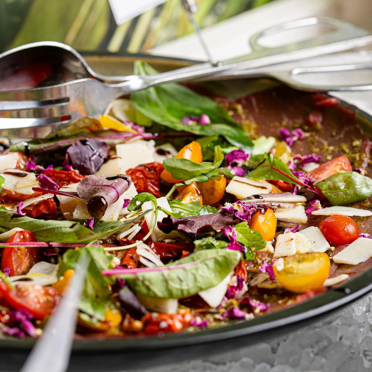Un grand saladier rempli d'une salade colorée contenant des tomates, des légumes verts, des radis, des dés de fromage et des graines, avec des ustensiles pour servir