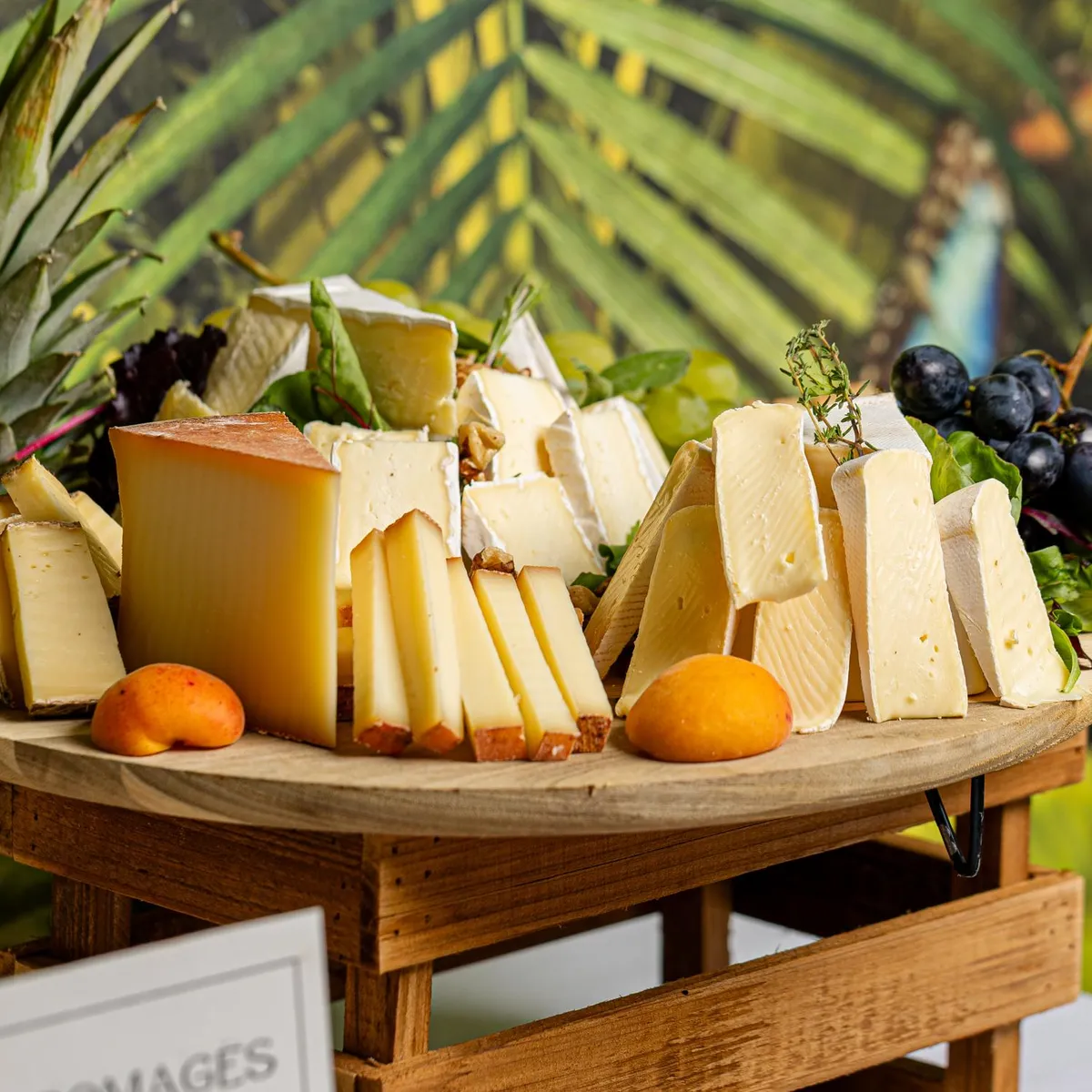 Un plateau en bois présente divers types de fromages, des fruits et des feuilles de basilic, entouré de plantes vertes