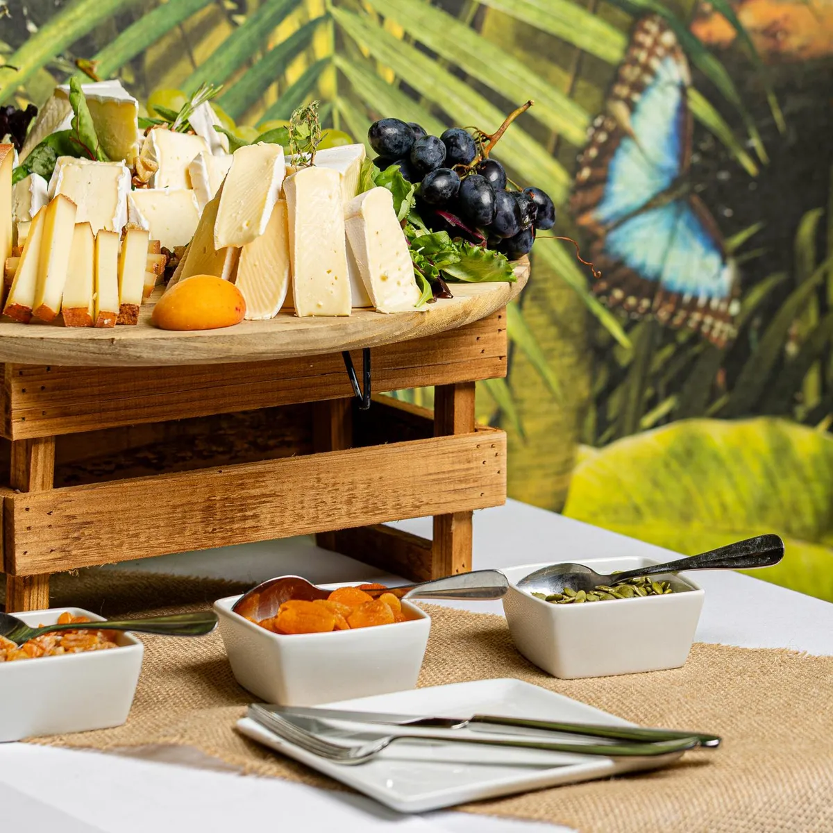 Un plateau de fromages et de fruits accompagné de bols de garnitures est présenté sur une table recouverte d'une nappe en jute avec une décoration tropicale en arrière-plan