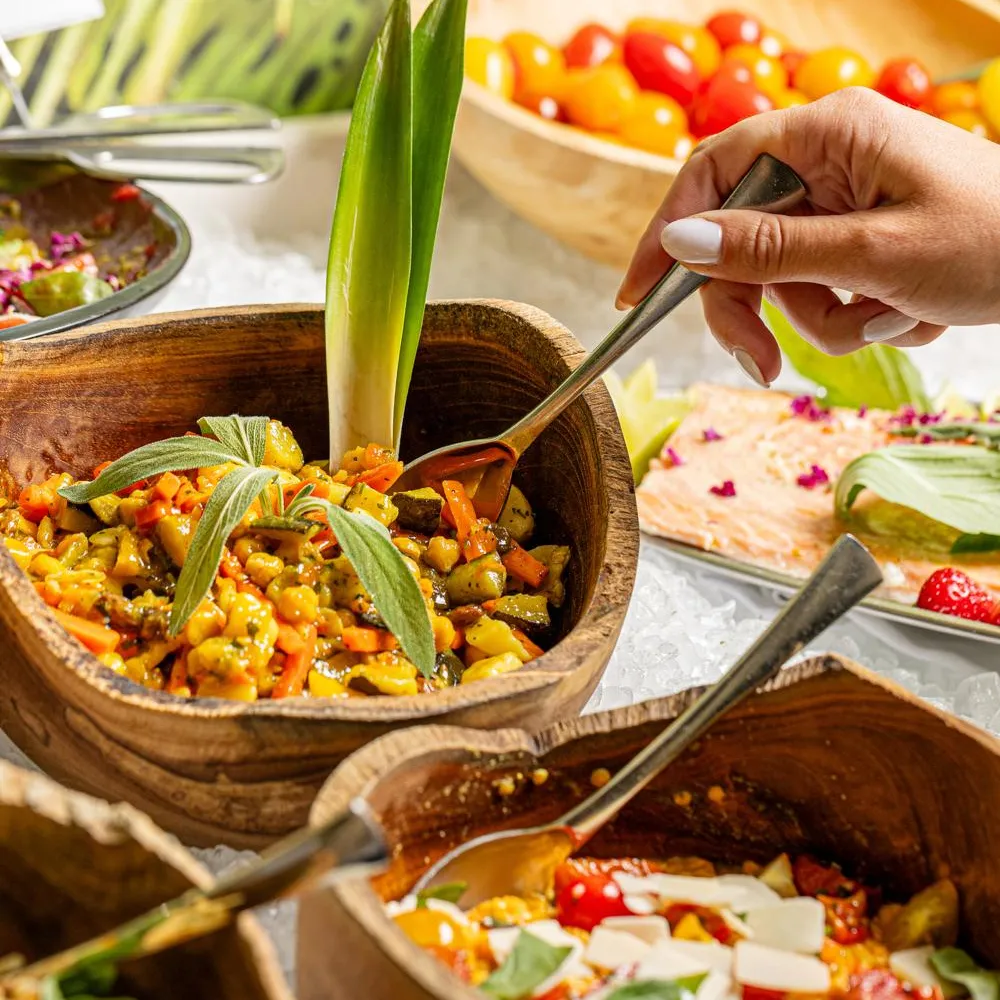 Une personne sert une préparation culinaire à base de légumes dans un bol en bois, entourée d'autres plats similaires