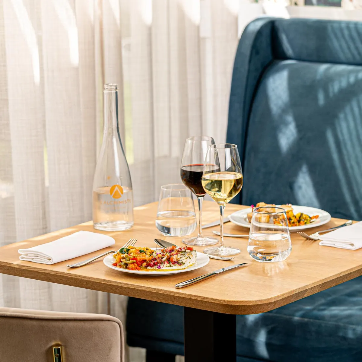 Table dressée avec deux assiettes de nourriture, deux verres de vin, deux verres d'eau, une bouteille d'eau et des couverts, devant une banquette bleue et une chaise beige