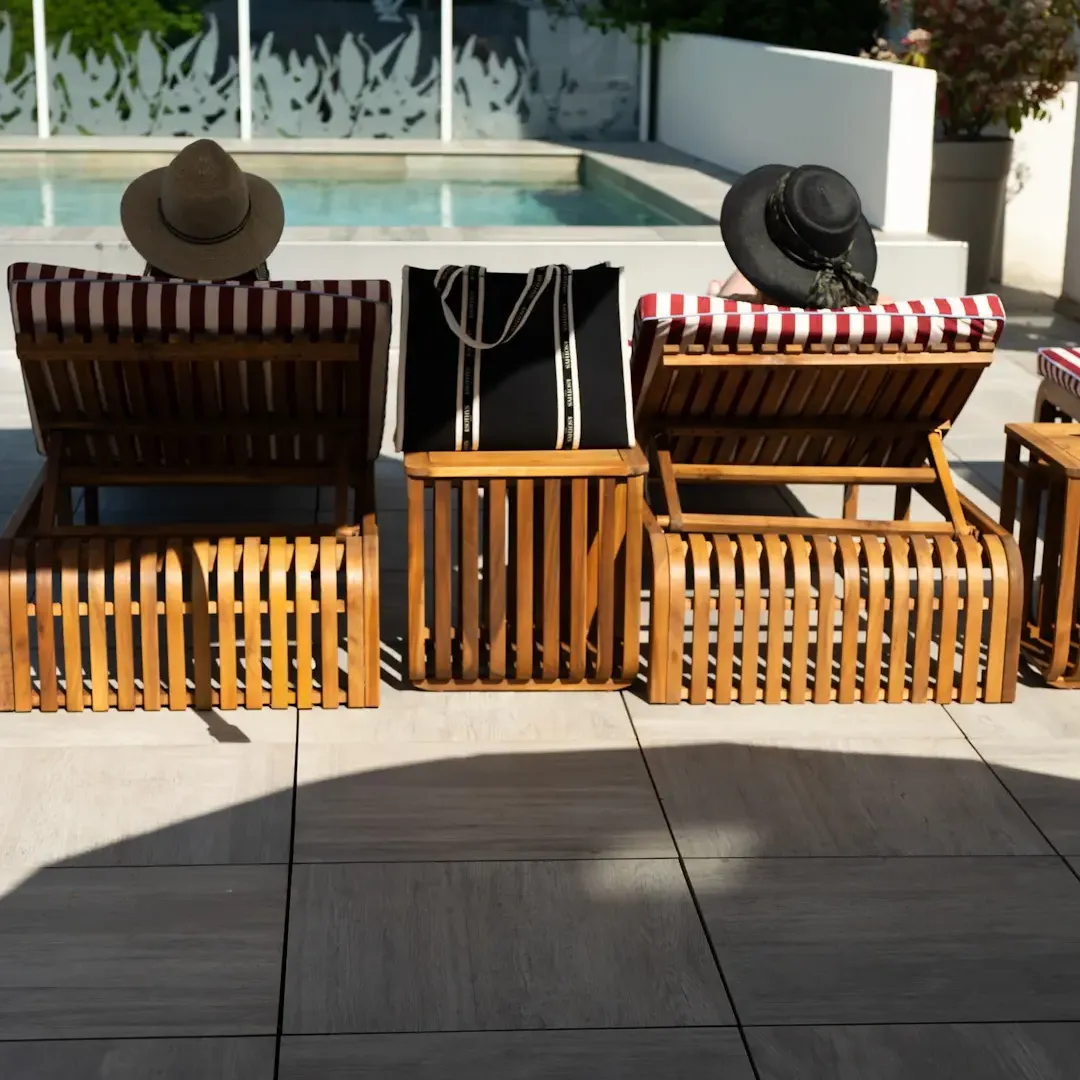 Two wooden deck chairs with red and white striped cushions, a black bag placed on one of them, in front of a pool with a metal railing and potted plants.