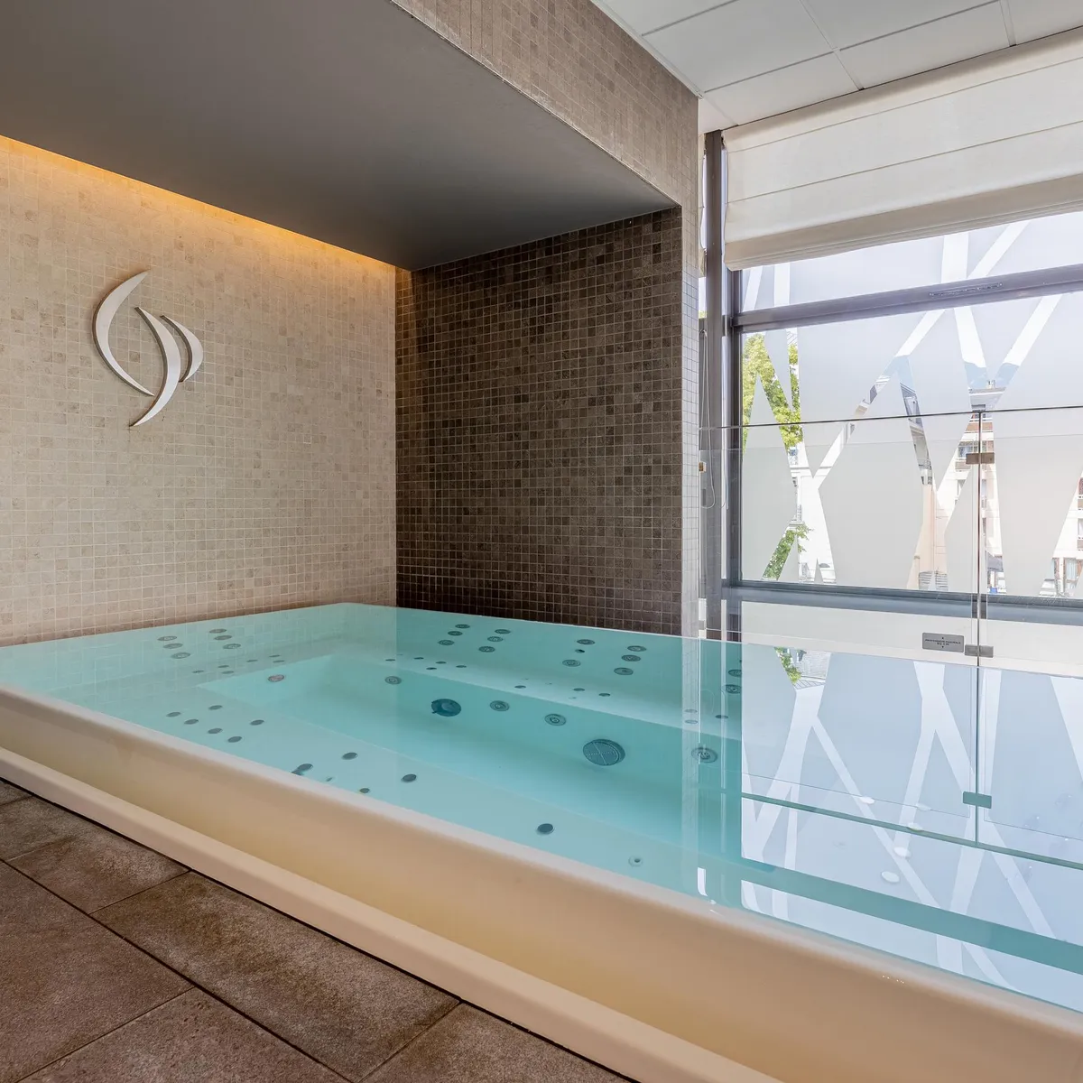 Piscine intérieure avec des jets d'eau, entourée de carreaux et de grandes baies vitrées donnant sur l'extérieur