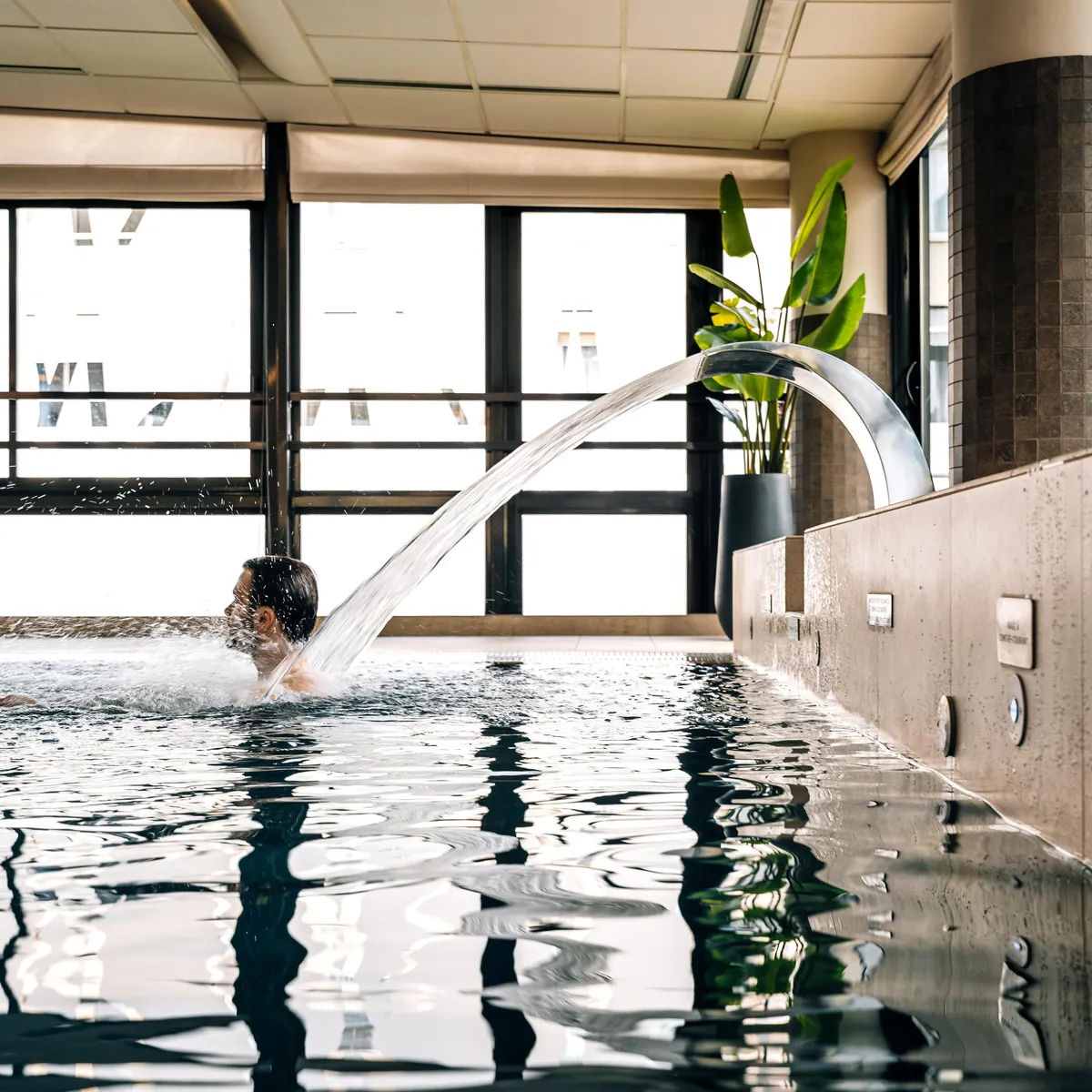 Une personne nage sous un jet d'eau dans une piscine intérieure avec de grandes fenêtres et une plante verte à proximité
