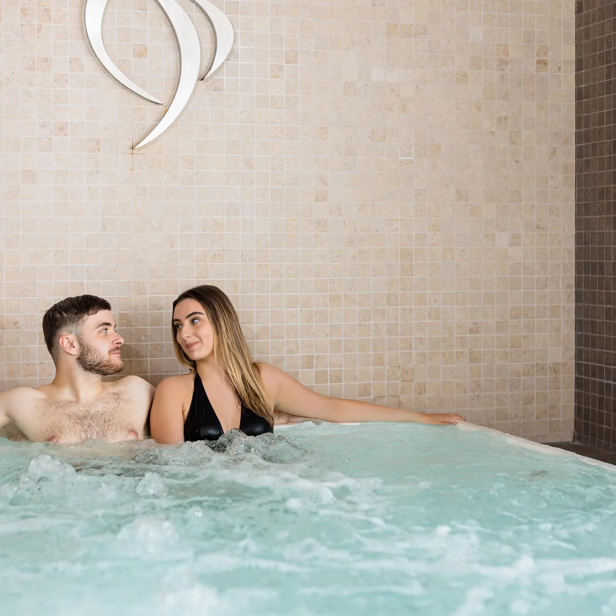 Un homme et une femme sont assis dans un jacuzzi avec des bulles, devant un mur carrelé beige avec un symbole blanc en forme de vague
