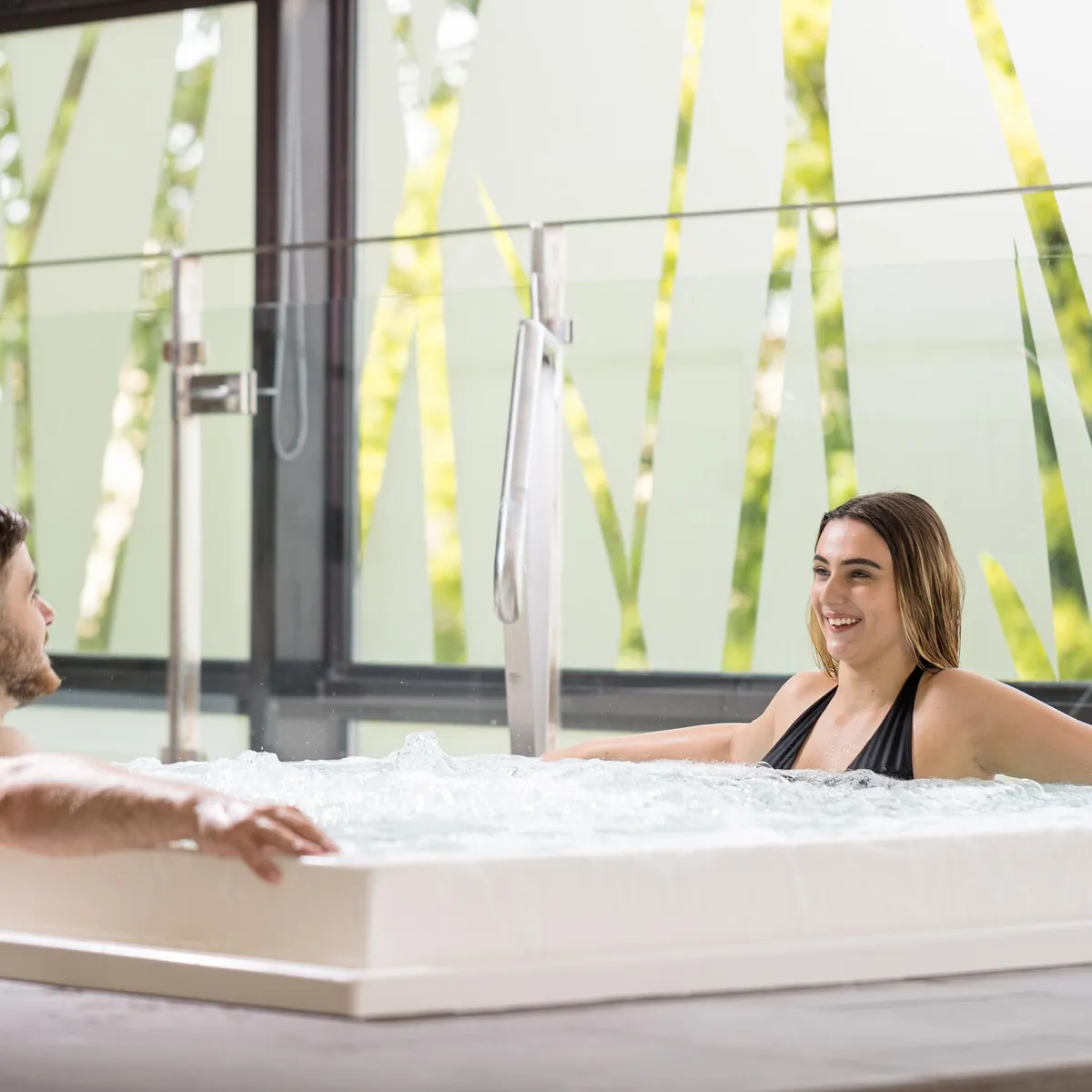 Un homme et une femme sont assis dans un jacuzzi, entourés de grandes fenêtres avec vue sur des plantes vertes