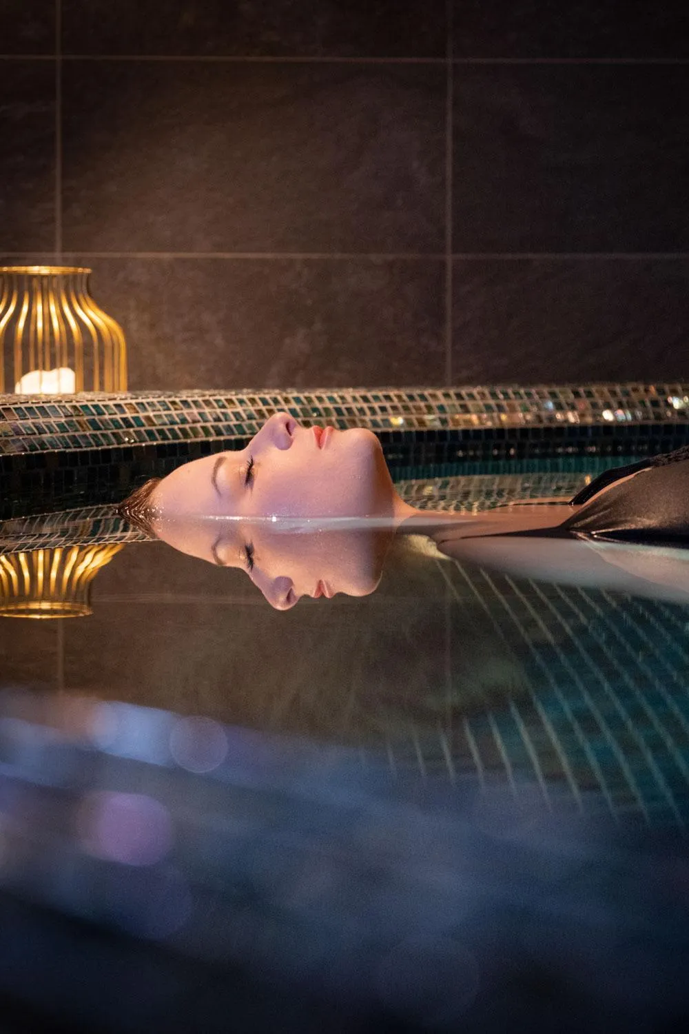 Une personne est allongée dans une piscine d'eau chaude, la tête reposant sur le bord, dans une ambiance tamisée avec des lumières et des carreaux visibles