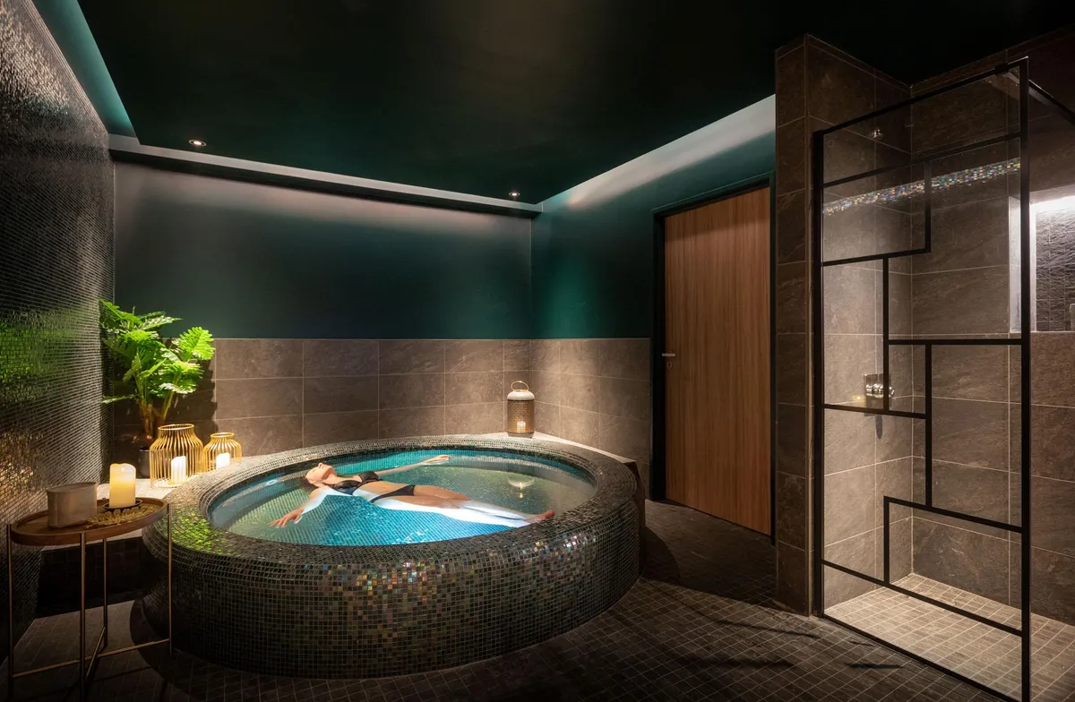 Eine Person entspannt sich in einem runden Whirlpool, der von Mosaiken umgeben ist, in einem Raum mit dunklen Wänden und gedimmtem Licht.