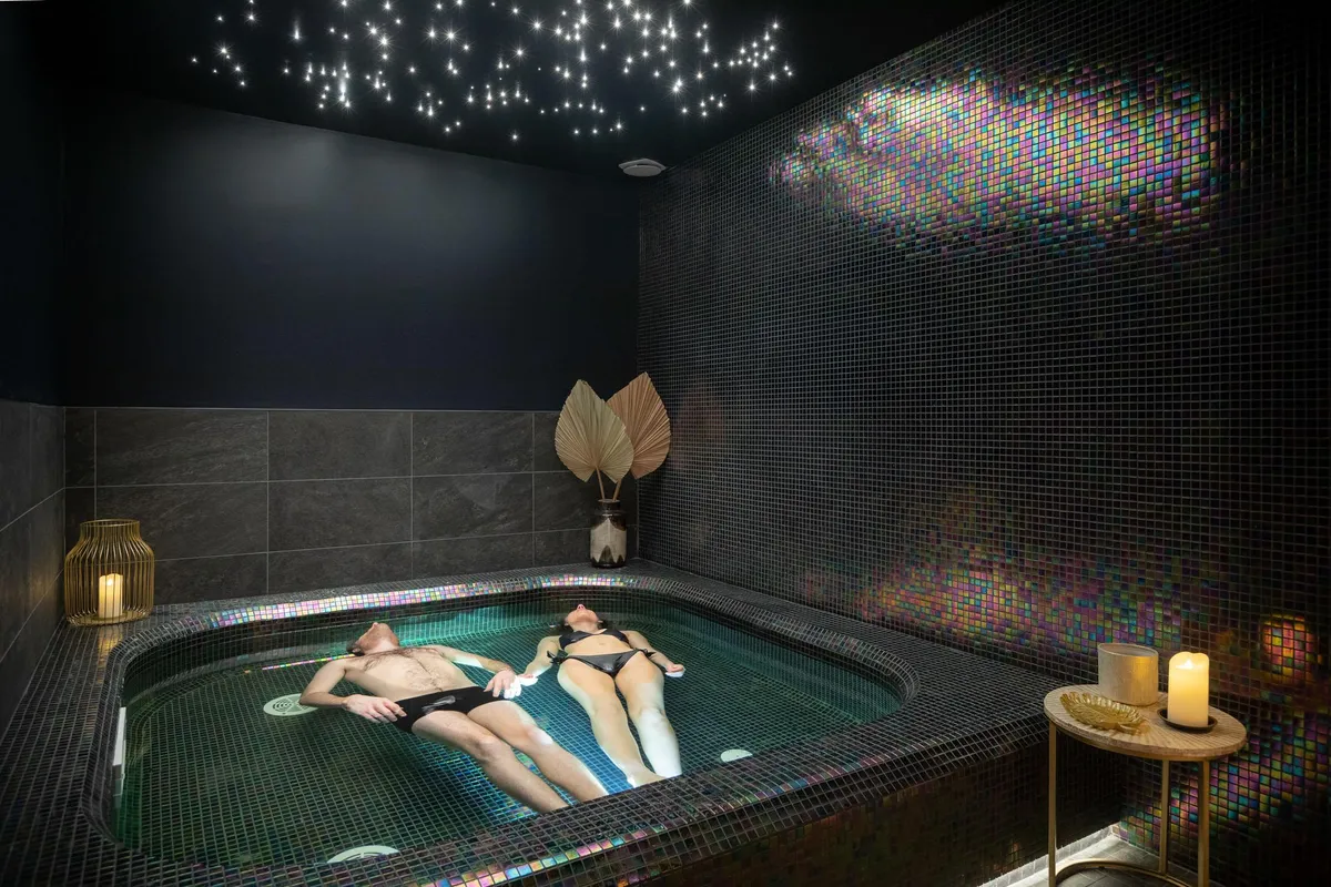 Zwei Personen liegen in einem Whirlpool, umgeben von bunten Mosaiken und Kerzen, mit einem Sternenhimmel an der Decke.