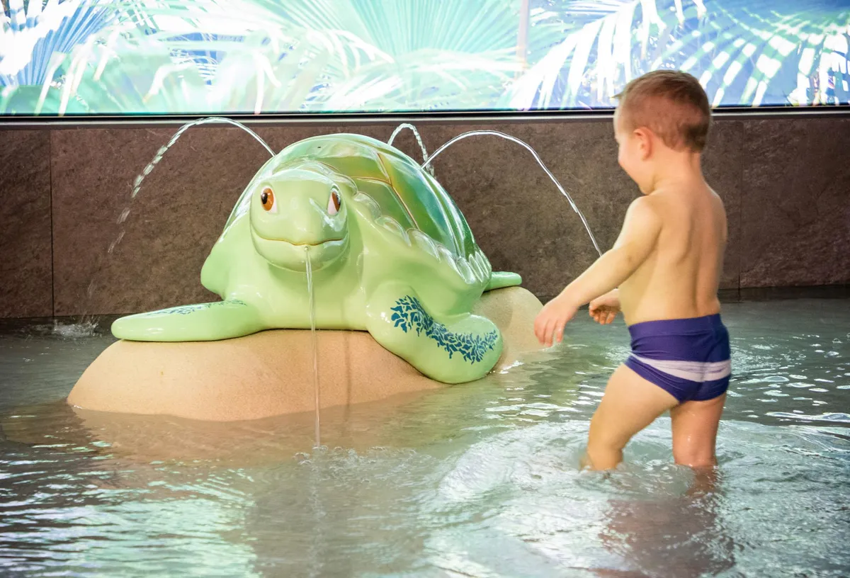 Enfant en maillot de bain bleu jouant avec une sculpture de tortue d'où s'écoule de l'eau dans une piscine peu profonde
