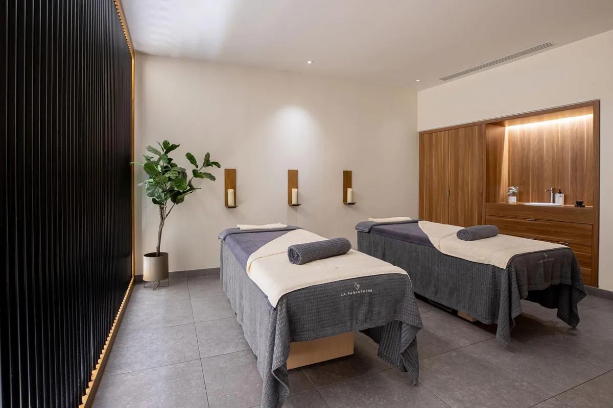 Deux tables de massage avec serviettes grises et blanches, une plante verte et des appliques murales dans une pièce aux murs beiges et au sol gris
