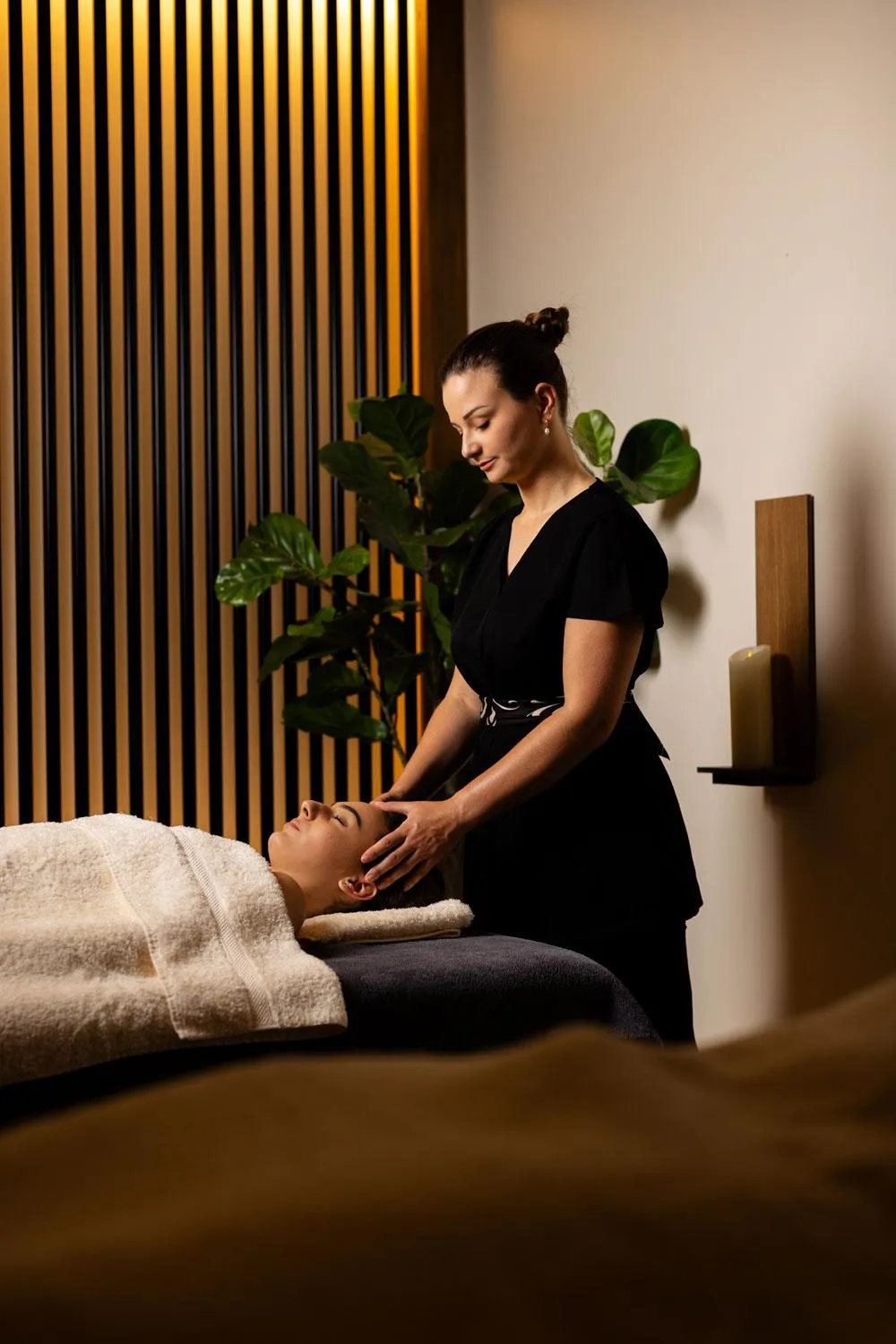 Une femme en tenue noire pratique un massage sur une personne allongée sur un lit de massage recouvert de serviettes