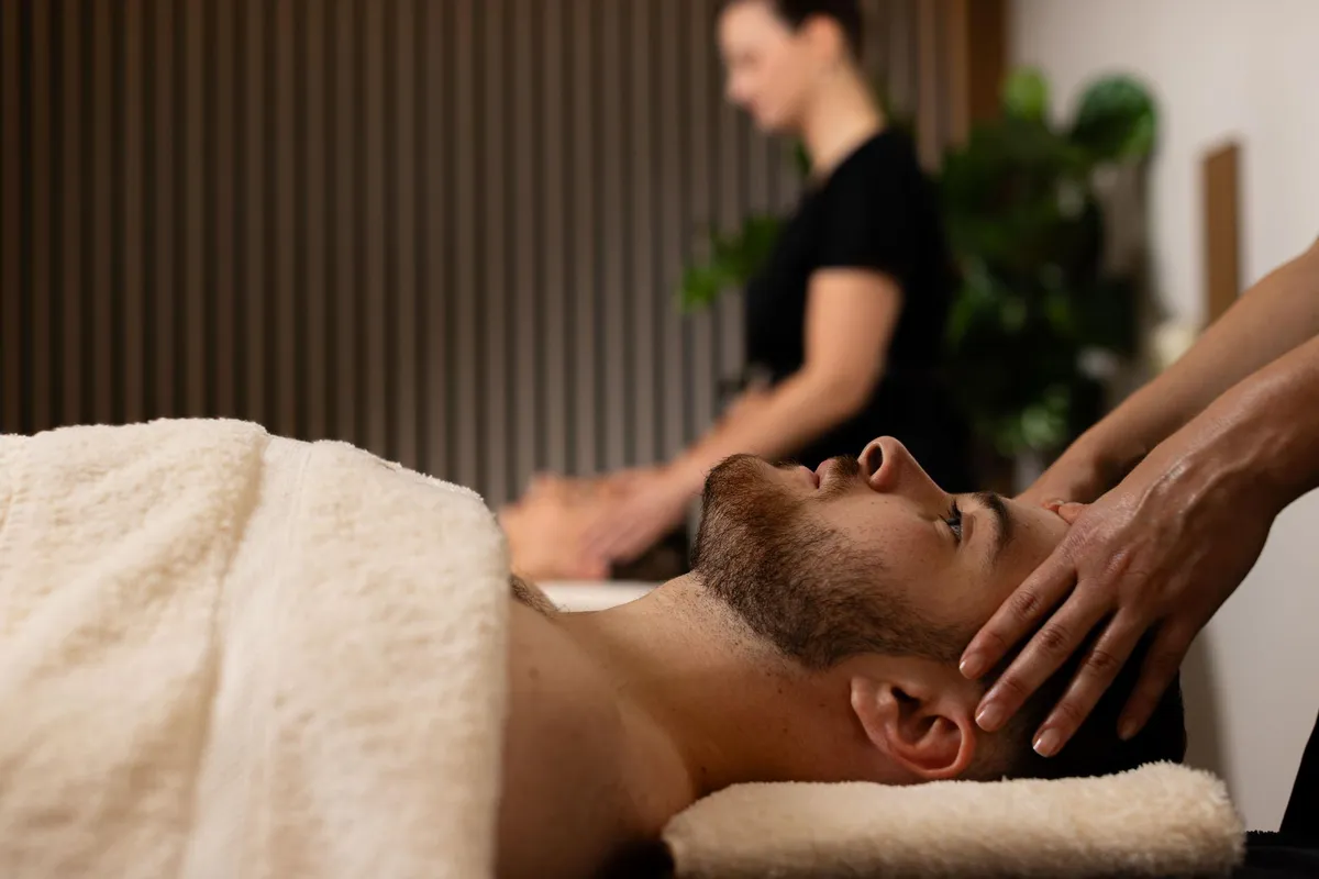 Un homme allongé sur le ventre reçoit un massage de la nuque par un professionnel, dans un cadre intérieur avec des plantes en arrière-plan