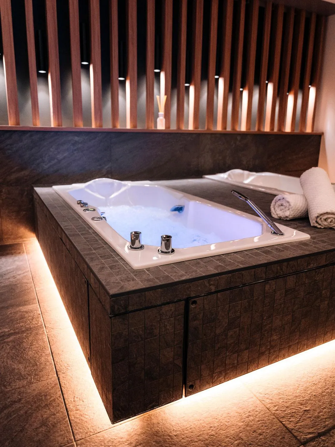 Jacuzzi filled with water and LED lights under the edge, surrounded by dark tiles and white towels.