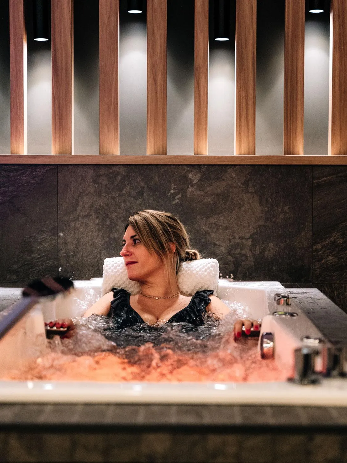 Une femme est allongée dans un bain à remous avec un repose-tête en fourrure, entourée de bulles, dans un cadre intérieur avec des colonnes en bois