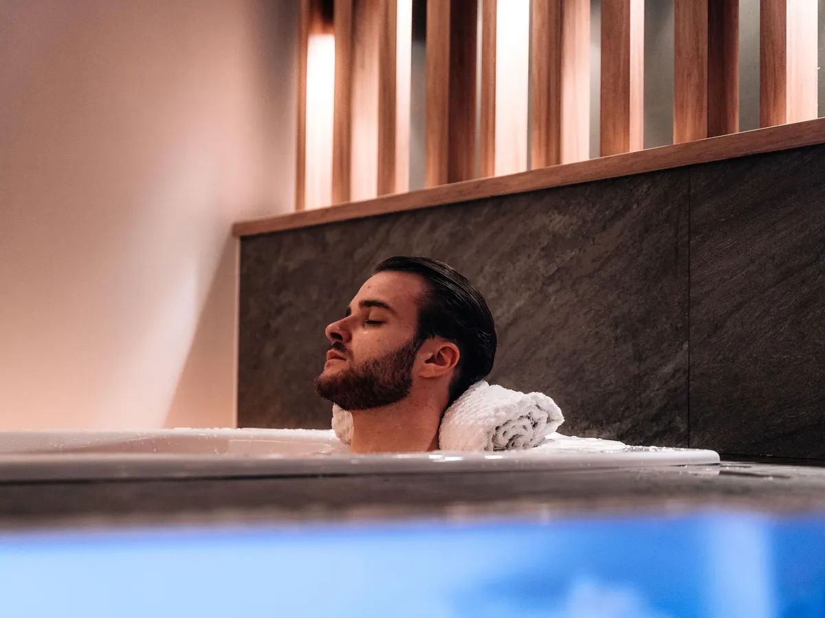 A bearded man is lying in a bath with a white towel over his shoulders, eyes closed.