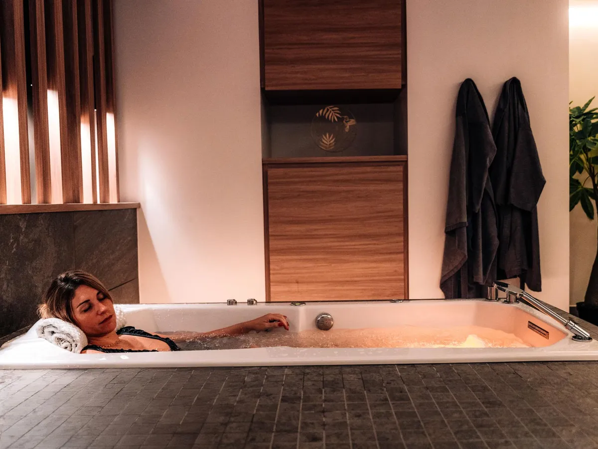 A person is lying in a bathtub filled with water, with towels placed on the edge and robes hanging nearby.