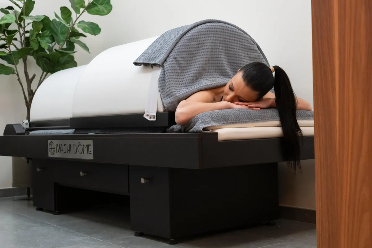 Une personne allongée sur un lit de massage recouvert d'une couverture grise dans une pièce avec une plante verte