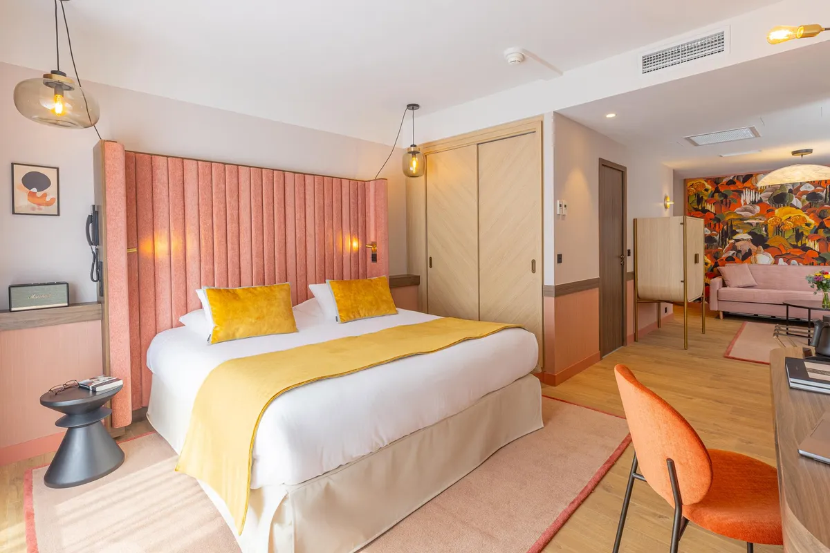 Chambre d'hôtel avec un lit double aux draps blancs et une couverture jaune, une tête de lit rose, une table de chevet avec des objets dessus, une lampe suspendue, un fauteuil orange et une porte ouverte sur un salon avec un canapé et une peinture murale