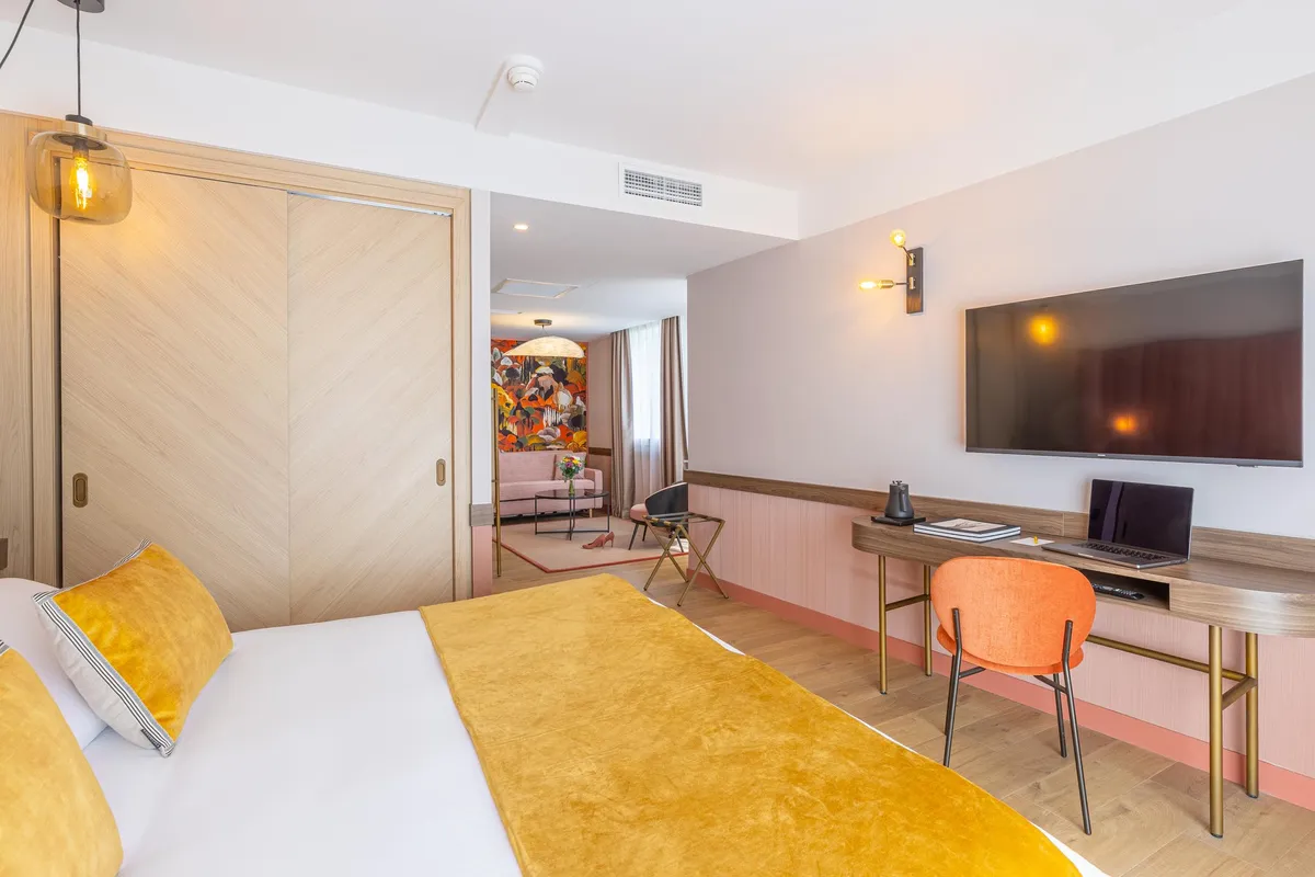 Chambre d'hôtel moderne avec lit, bureau, télévision et espace salon visible au fond