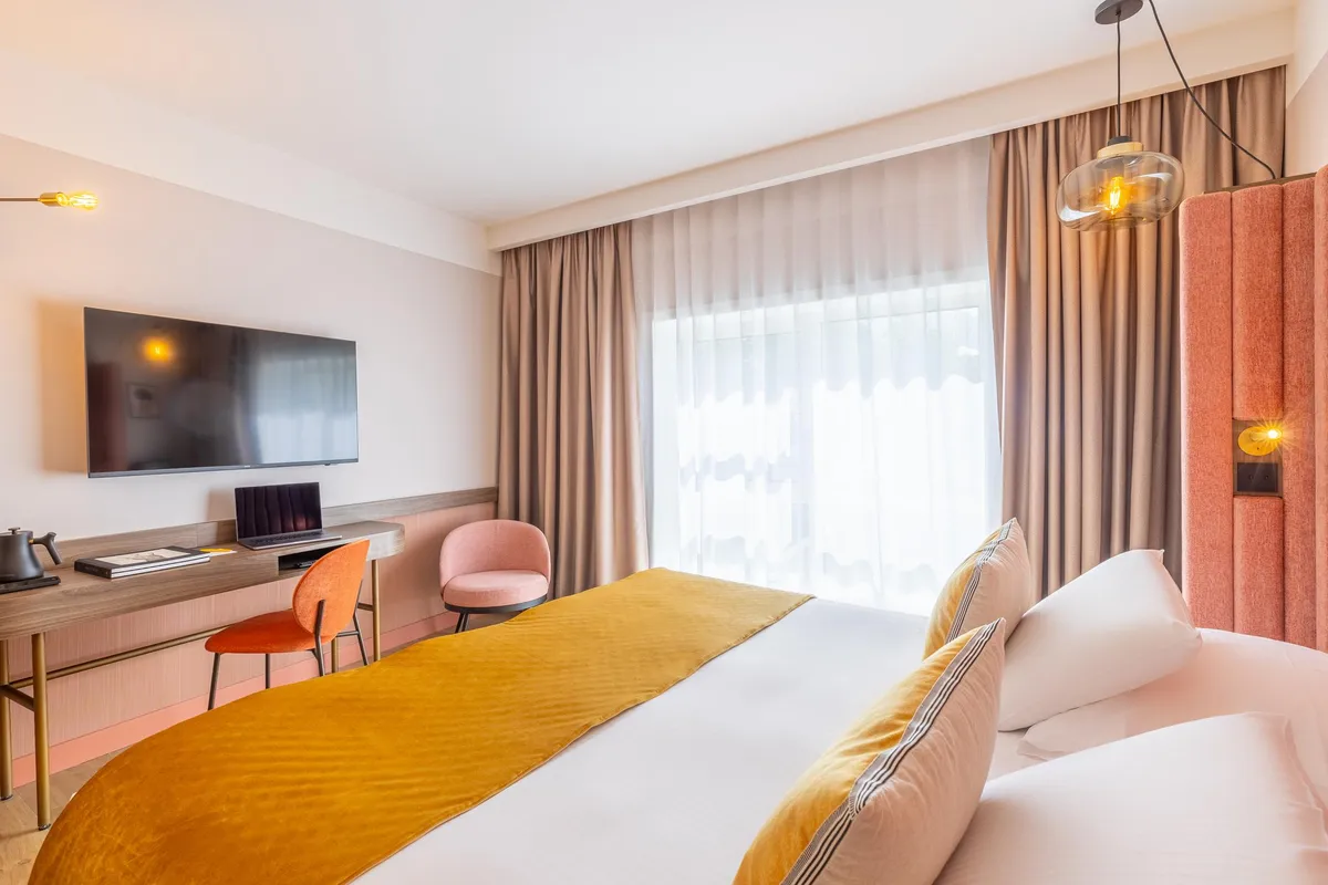 Modern hotel room with a double bed, a TV, a desk, and a large window with curtains.