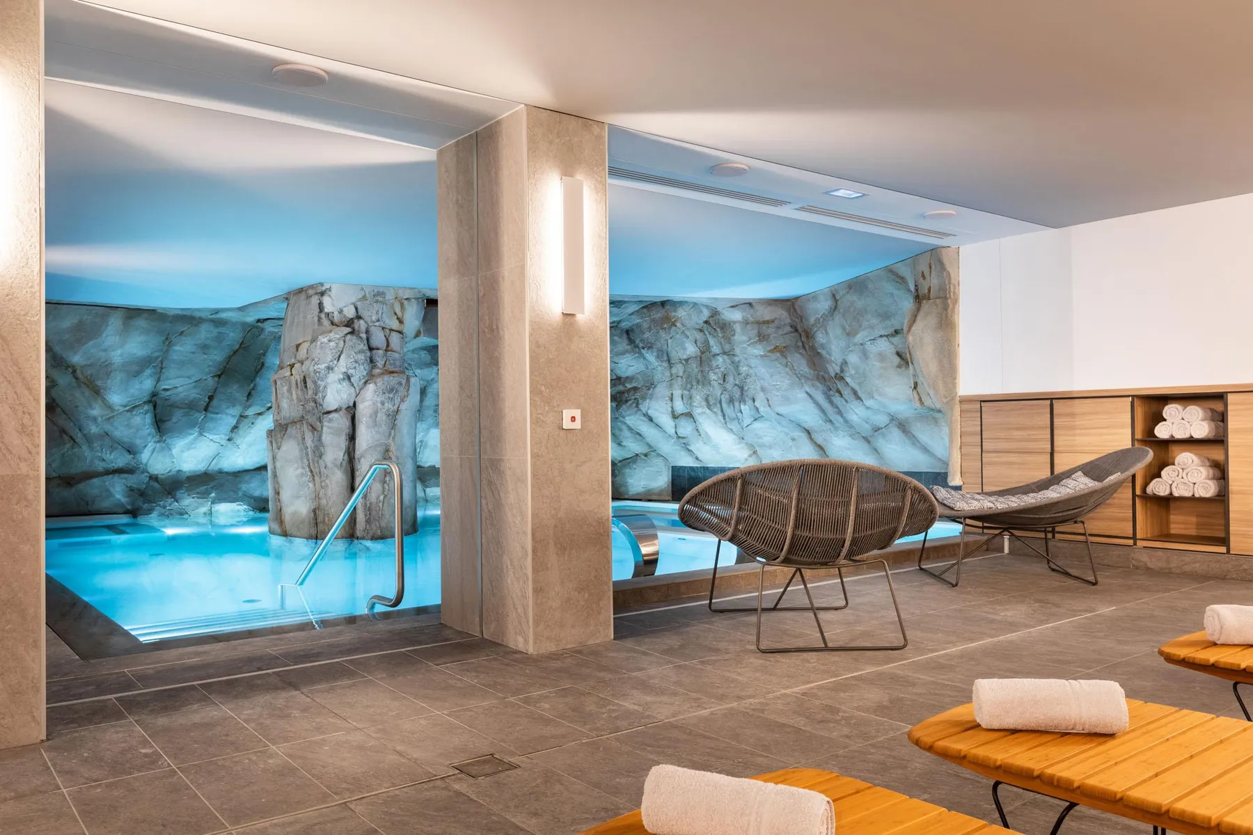 Salle de relaxation avec des chaises longues, des serviettes et une vue sur une piscine à travers une grande vitre décorée de rochers